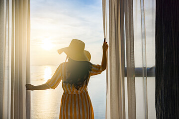 Rear back view woman opening curtains, relaxing and looking beautiful seascape panorama with rays...