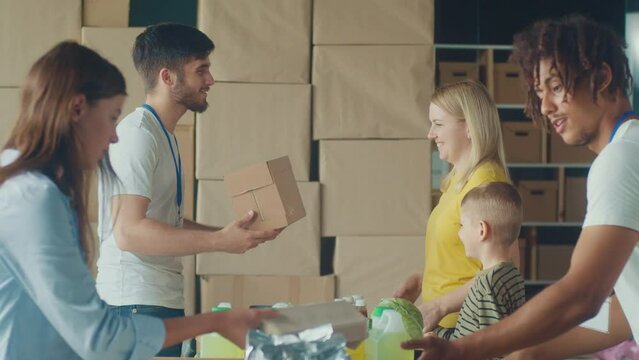 Friendly Volunteer Giving Donation Box With Food to Woman and Her Son at Distribution or Refugee Assistance Center. The Concept of Social Aid. Internally Displaced Persons