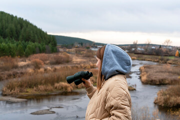 young woman ecologist in beige jacket and hood with binoculars watching swimming birds on river and...
