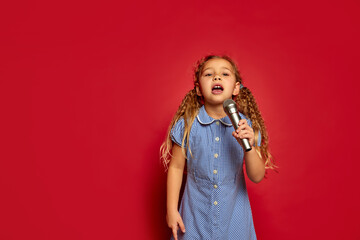 Cute little kid girl isolated on red studio background. Human emotions, facial expression. Concept of childhood, children, lifestyle. Copy space for ad. She listen music, sing song in microphone