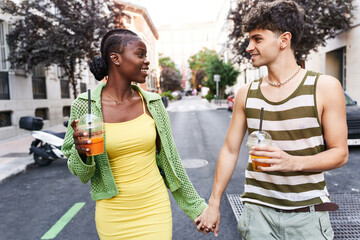 Loving young multiethnic couple holding hands and drinking juices while walking on street