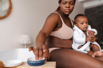 Obraz na płótnie Canvas Young african woman applying skin cream while holding with her baby