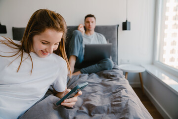 Young white woman holding cellphone while man using laptop in bed