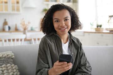 Smiling mixed race girl holding smartphone with modern apps, looking at camera, sits on sofa at home