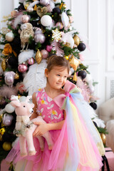 Smiling beautiful girl in pink elegant dress with a deer toy and new year tree on the background.