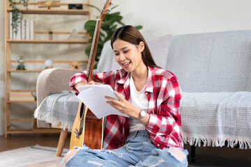 Young woman is compose song and writing lyrics on notebook while putting guitar on the legs and...