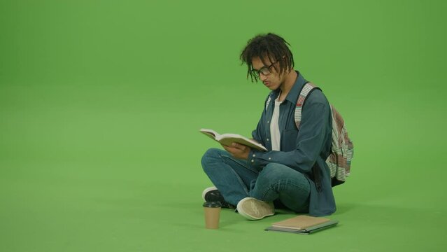 Young Multicultural Man in Glasses and Denim Shirt Looking to Book,Turn Pages of Book and Smiling. Portrait of Handsome Guy Sitting with Books,Notes and Coffee on Green Screen Background