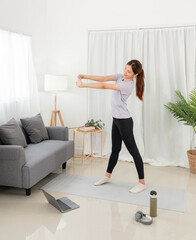 Young woman is taking yoga lesson online on laptop and workout yoga exercise while standing to stretching