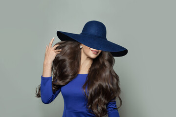 Young beautiful brunette woman in blue dress and wide broad brim hat posing on white background