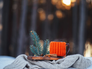 A mug of sea buckthorn tea in a knitted orange jacket in the winter forest in the snow. Steam from...