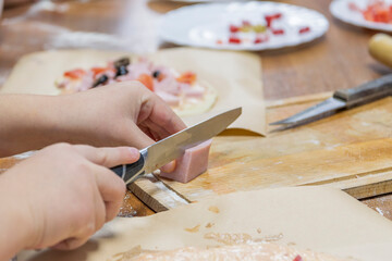 Cropped hands of child on cutting board cut piece of sausage for cooking pizza with kitchen knife