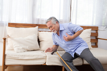 Senior Asian man falling onto couch and holding chest and dropping cane because of a painful heart...