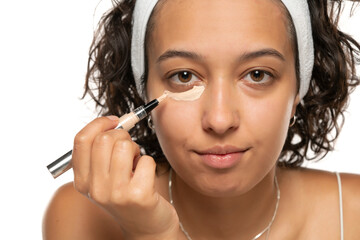 Young etnic woman applyes concealer under her eyes on white background