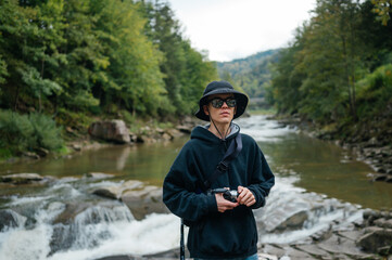 Portrait of a handsome male tourist in stylish casual clothes and sunglasses posing for the camera against the background of a mountain river and forest, holding an old digital camera