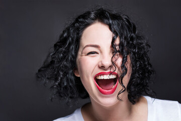 Young girl laughing contagiously with her mouth open. Beautiful bright curly brunette in a white tank top with red lipstick. Black background. Close-up.