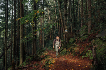 Male tourist is hiking alone in the mountains with a stick in his hands, walking along a path in the forest and looking away with a serious face. Active recreation in the mountains.