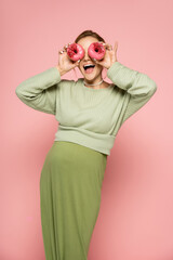 Excited pregnant woman covering face with donuts on pink background.