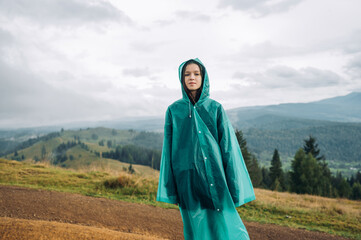 Attractive woman hiker in raincoat stands in mountains in bad weather during a hike and looks at the camera against the background of beautiful views.