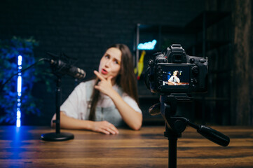 Funny woman sitting at a table and recording videos on camera with a funny face in a professional studio. Focus on the camera.