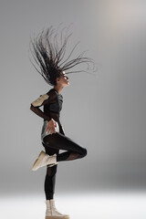 side view of fit woman with dreadlocks wearing tight jumpsuit and silver shorts on grey background.