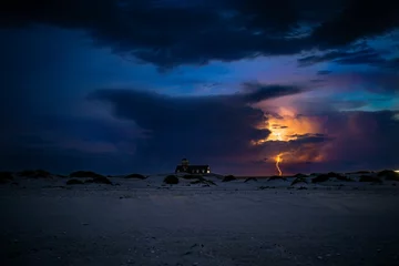 Fotobehang Big sandy beach and a lighthouse with lightning in the dark blue sky in the background © Lumosphotos/Wirestock Creators
