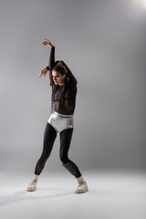 full length of brunette woman in futuristic clothes posing with hands above head on grey background.
