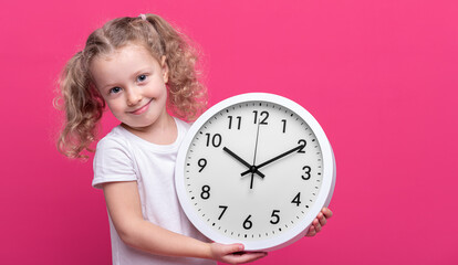 A little girl is holding a white clock on a pink background.