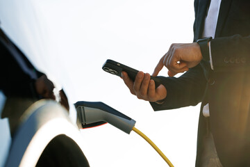 Businessman charging electric car at outdoor charging station. Unrecognizable man plugging electric...
