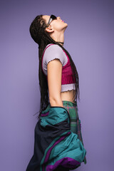 side view of nineties style woman in sunglasses posing with raised head isolated on purple.