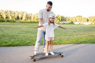 father help his daughter stand on skateboard. Dad teaching her to ride in park.