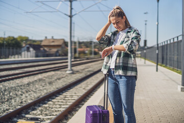 Upset woman looking at her clock while standing with suitcase  on a train station.