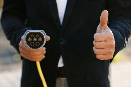Businessman hand showing thumbs up holding power type 2 ccs cable supply plugged at electric car charging station. Man standing near electric charging station and showing thumb up