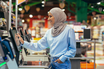 A woman buyer in a supermarket in a hijab pays for goods at a self-service checkout, convenient service for customers. - obrazy, fototapety, plakaty
