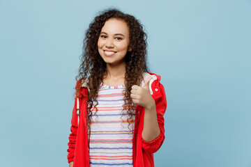 Young smiling happy satisfied woman of African American ethnicity wear red raincoat showing thumb up like gesture isolated on plain pastel light blue cyan background. Wet fall weather season concept.