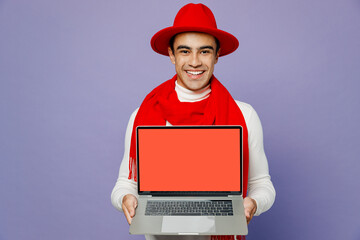 Young middle eastern IT man wear white turtleneck red hat scarf hold use work on laptop pc computer with blank screen area isolated on plain pastel light purple background. People lifestyle concept.