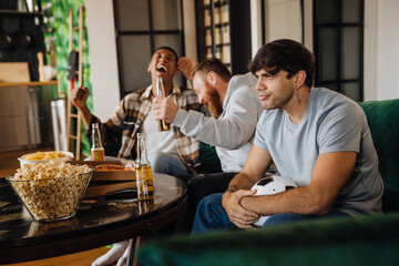 Displeased man watching sports match while his friends celebrating victory in living room