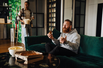 Two male friends holding beer and pizza while watching sport match in living room