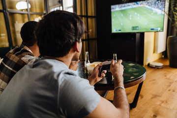 Back view of young male friends playing football video game with gamepads at home