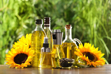 Many different bottles with cooking oil, sunflower seeds and flowers on wooden table against blurred green background