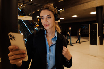 Young woman in headphones looking at mobile phone screen while standing by the escalator