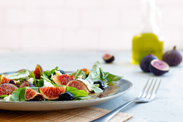 Close-up of fresh healthy summer salad with figs, white cheese, arugula, black olives and sauce on ceramic plate and fork on white background. Selective focus. Blurred background.