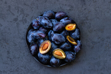 Fresh ripe blue whole and cut plums on plate on dark gray background. Low key photo. Top down view. Close-up. Flat lay composition. Copy space.