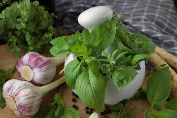 Mortar with different fresh herbs near garlic, horseradish roots and black peppercorns on wooden table, closeup