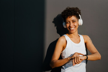 Fototapeta na wymiar Black woman in headphones using smartwatch while standing by wall