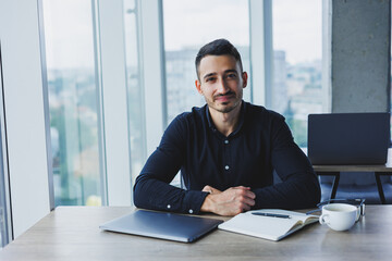 A successful businessman man of Caucasian appearance in a black shirt is sitting at a desk using a laptop in a modern office. Working atmosphere in the office.
