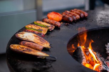 Process of cooking meat sausages, zucchini, eggplant, tomato on grill, brazier at outdoor summer local food market - close up. Gastronomy, cookery and street food concept