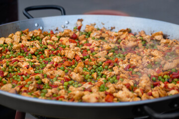 Process of cooking chicken meat pieces, peppers, peas for Spanish paella in huge wok at summer outdoor food market - close up, selective focus. Outdoor cooking, gastronomy and street food concept