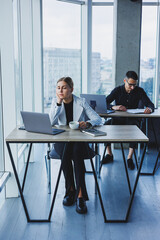 A female manager is sitting at a desk using a laptop in a modern office against the background of a colleague. Working atmosphere in the office.