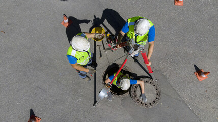 Maintenance and washing of a cesspool through the access of a road manhole. Skilled workers at work lower themselves underground using a pulley - obrazy, fototapety, plakaty
