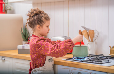 A little girl in the kitchen in an apron cooks something in a saucepan on a gas stove.